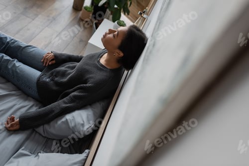 Preview: High angle view of sad multiracial woman in knitted sweater leaning on wall while sitting on bed at