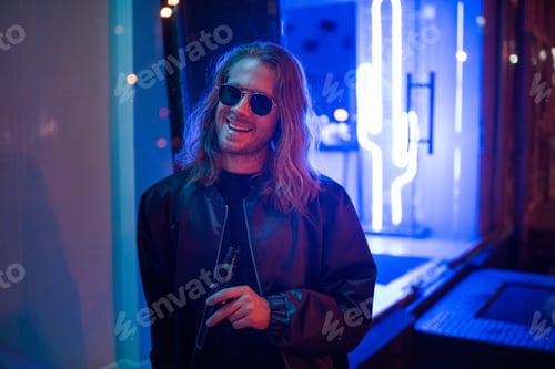 Preview: happy young man in leather jacket and sunglasses with bottle of beer on street at night under blue