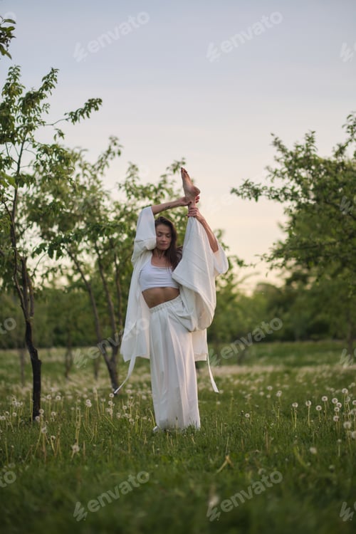 Preview: A woman dressed in white stretches her leg upwards in a grassy meadow, framed by trees. This scene