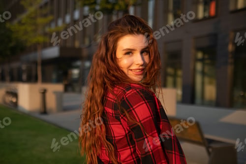 Preview: Young woman with a radiant smile and red hair stands against the backdrop of a business center.