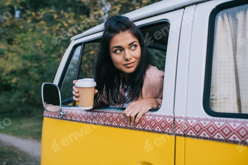 Preview: beautiful traveler with disposable cup of coffee sitting in retro minivan
