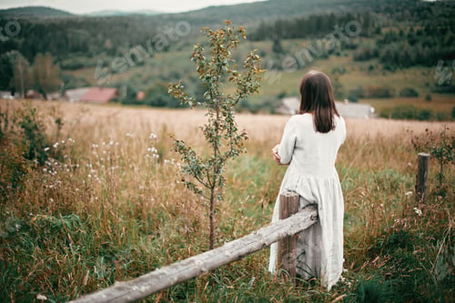Preview: Stylish girl in linen dress sitting on aged wooden fence among herbs and wildflowers