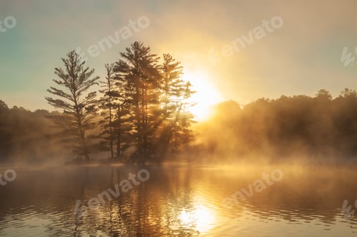 Preview: Small lake and island with sunburst shrouded in fog on a summer sunrise in Minnesota