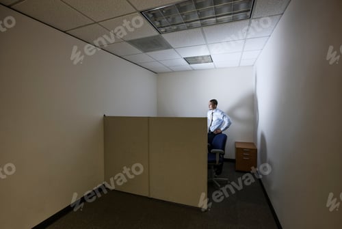 Preview: A businessman lit up by a computer screen in his small corner office cubicle.