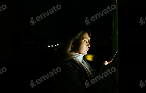 Preview: Young caucasian woman using mobile phone outdoor in the evening, walking on city streets.