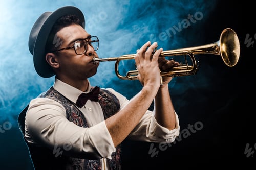 Preview: male musician in hat and eyeglasses playing on trumpet on stage with dramatic lighting and smoke
