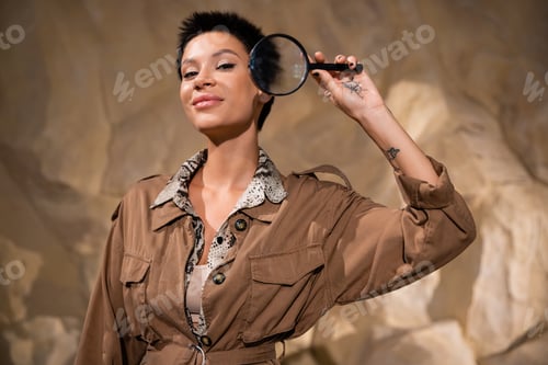Preview: happy archaeologist with tattoo and short hair holding magnifying glass in cave