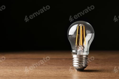 Preview: Vintage filament lamp bulb on wooden table against black background. Space for text