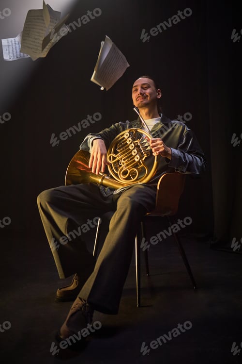 Preview: Smiling French Horn Player Sitting in Chair with Instrument in Black Studio