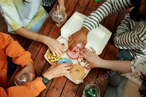 Preview: Teenagers Reaching for Donut from Takeaway Box Sitting at Table Outdoors