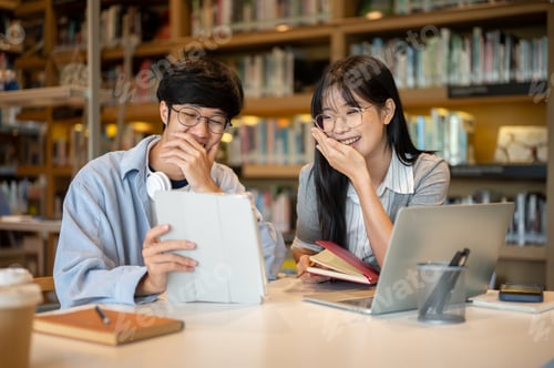 Preview: Glasses asian man student and woman friend looking and laughing at tablet at wooden table in library
