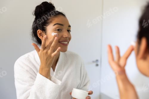 Preview: Happy biracial woman in bathrobe applying cream on her face in sunny bathroom