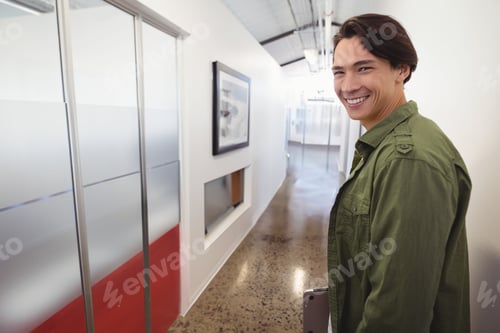 Preview: Asian man smiling and holding closed laptop while standing in modern office corridor, copy space