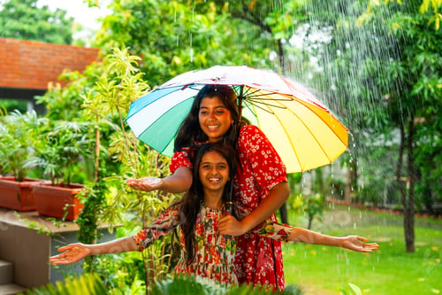Preview: Two Indian girls playfully enjoy monsoon rain under umbrella, sharing joyful outdoor moment