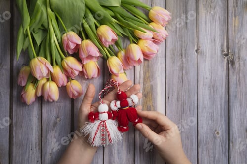 Preview: Martenitsa, Martisor in hands against the background of a bouquet of pink tulips on a wooden table
