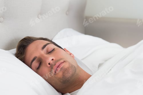 Preview: Man lying under white bedding with head resting on white pillow against beige headboard in bedroom