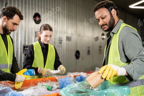 Preview: Bearded indian worker in safety vest and protective gloves putting paper cup in plastic bag while