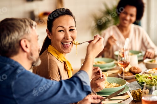 Preview: Happy Asian woman having fun while husband is feeding her at dining table.
