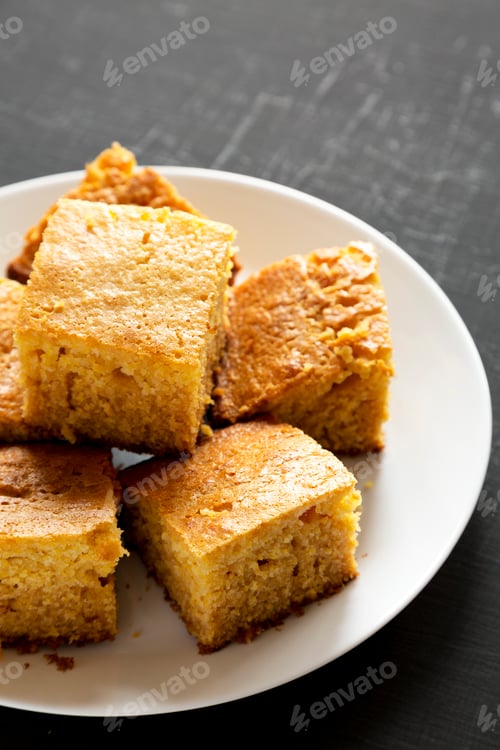 Preview: Homemade Sliced Cornbread Ready to Eat on a white plate on a black background, low angle view.