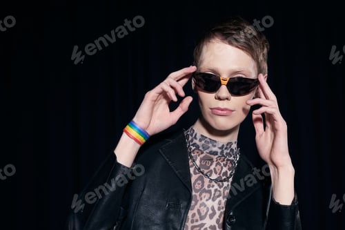 Preview: Stylish homosexual man in bracelet with lgbt flag touching sunglasses while posing during pride