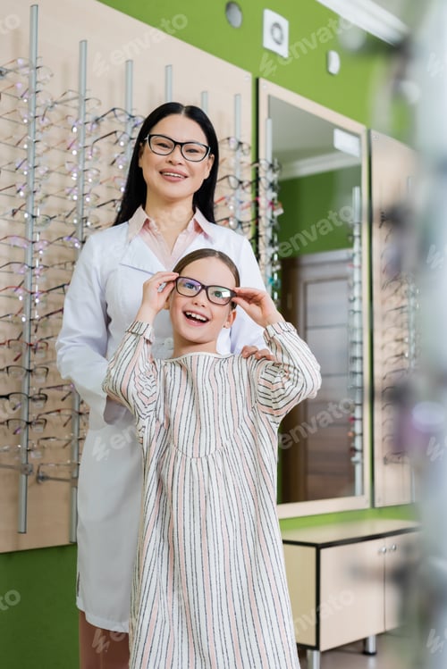 Preview: happy girl trying on eyeglasses near smiling asian oculist in optics shop on blurred foreground