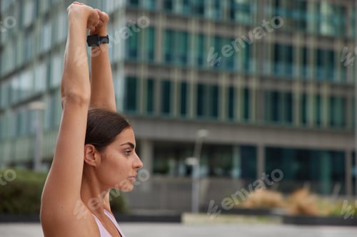Preview: Profile shot of sporty woman raises arms warms up before workout wears smartwatch focused into dista