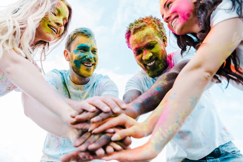 Preview: low angle view multicultural friends with holi paints on faces putting hands together