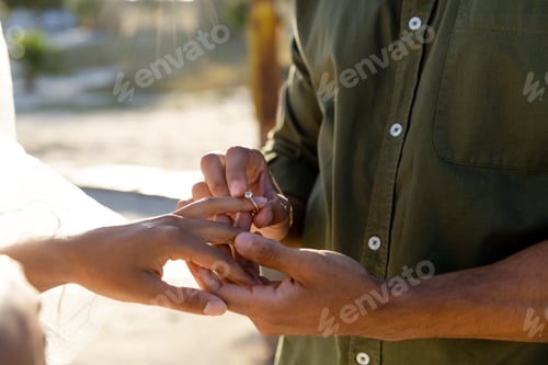 Preview: Midsection of caucasian young groom wearing wedding ring to bride at wedding ceremony on beach