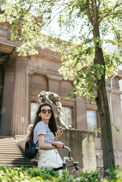 Preview: beautiful smiling girl in sunglasses standing with bicycle and using smartphone