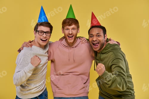 Preview: three multicultural cheerful friends in birthday hats hugging and looking at camera, birthday