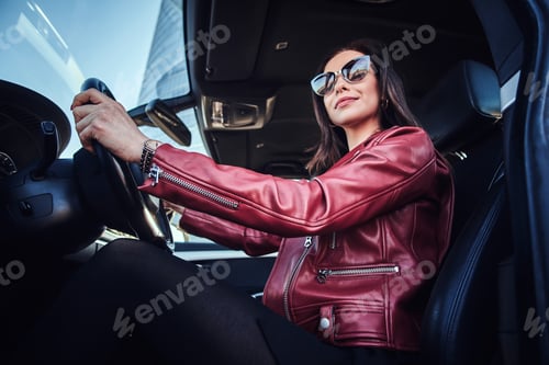 Preview: Attractive young woman in red jacket and sunglasses in driving her car