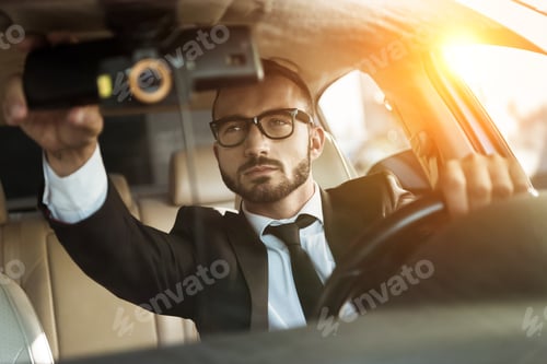 Preview: handsome driver in suit driving car and fixing mirror during sunset