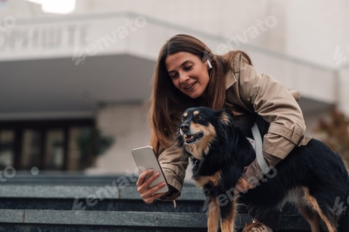 Preview: Smiling woman hugging her dog showing him something on smartphone screen while sitting on stairs