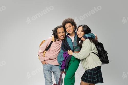 Preview: pleased and happy multiethnic students with backpacks smiling at camera and embracing isolated on