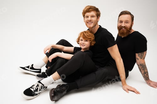 Preview: bearded tattooed man with redhead son and grandson smiling at camera while sitting on light grey