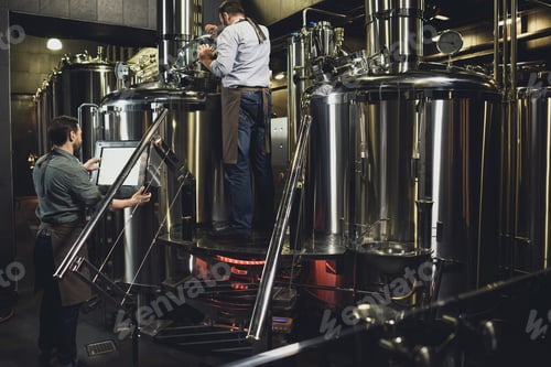 Preview: Male brewers in aprons working with tanks and industrial equipment at the brewery