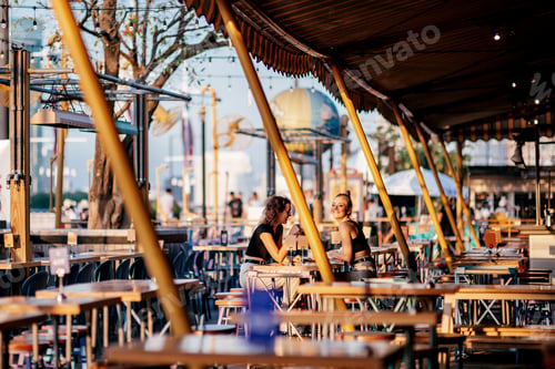 Preview: Vacay Mode. Girls sitting at riverside restaurant near Chao phraya river during sunset in the city