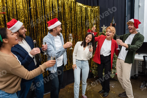Preview: Positive multicultural business people in santa hats holding champagne near tinsel in office