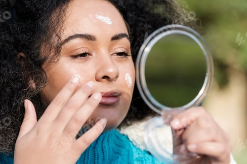 Preview: Front view of young woman applying moisturize in front of a mirror outdoors.