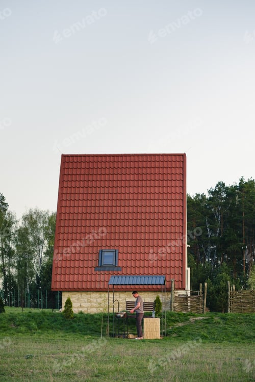 Preview: Man Prepares Food on Grill Near a House with Red Tiled Roof on Grassy Lawn