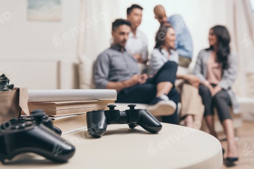 Preview: young casual multiethnic friends sitting on sofa with joysticks on foreground