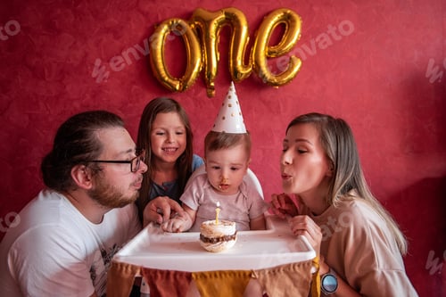 Preview: Mother father help blow out candle to one year old son on birthday cake. Happy family celebrating