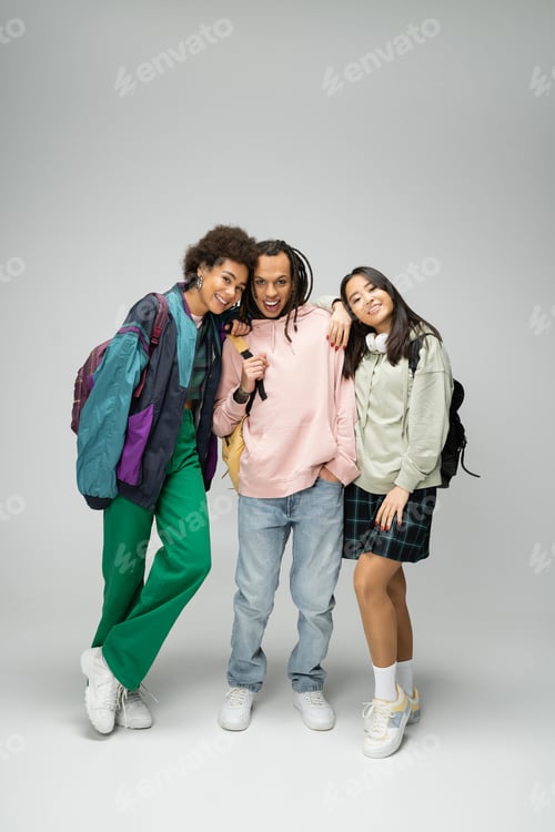Preview: full length of trendy student with dreadlocks posing near pretty interracial women on grey
