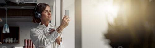 Preview: Smiling Business woman holding cup of coffee and using phone while standing near window at office