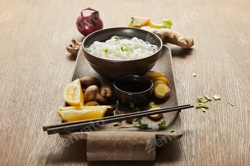 Preview: rice noodles in bowl near chopsticks, soy sauce, ginger root, lemon and vegetables on wooden tray