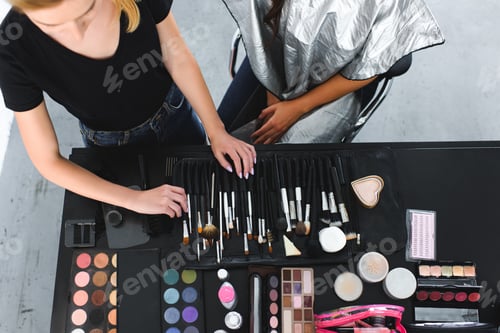 Preview: overhead view of makeup artist and woman covered with cloth at tabletop with makeup equipment
