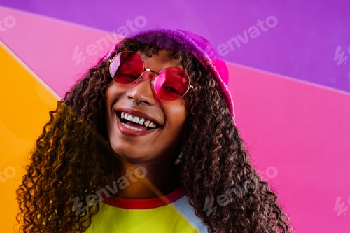 Preview: Young woman wearing pink sunglasses and beanie smiling on colorful background