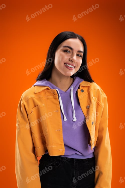 Preview: young woman in purple hoodie and stylish jacket smiling at camera isolated on orange