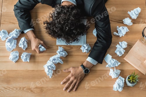 Preview: overhead view of tired businessman lying on table with crumpled papers