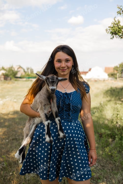 Preview: Young woman playing with cute baby goat standing near the barn